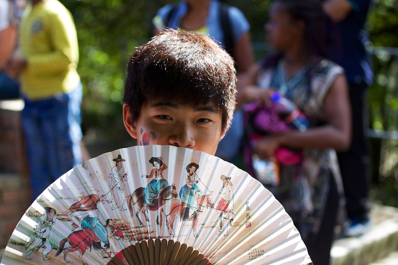 An Asian looking boy is covering a part of his face with a fan
