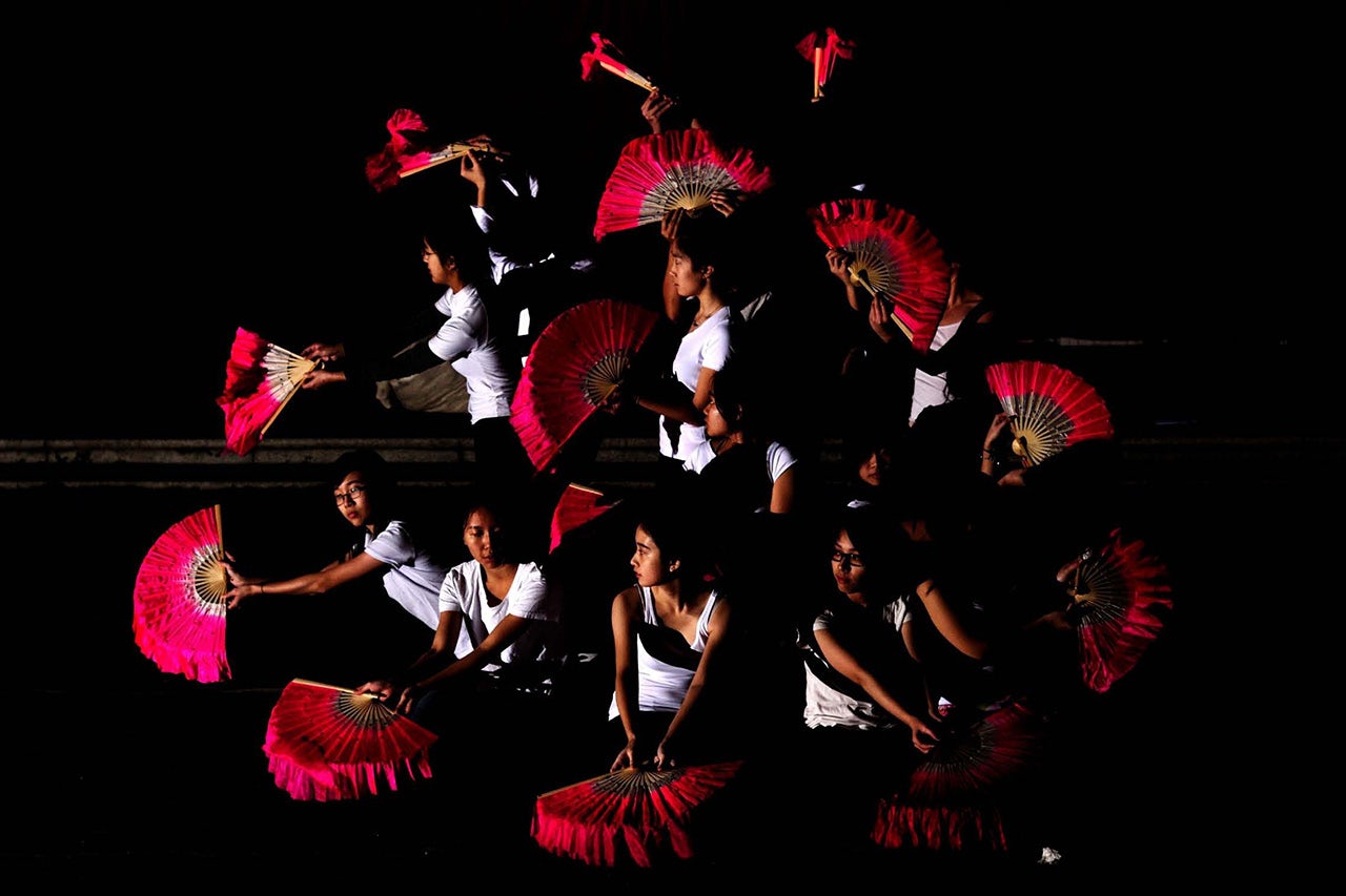 A group of dancers with red fans perform in front of a black background