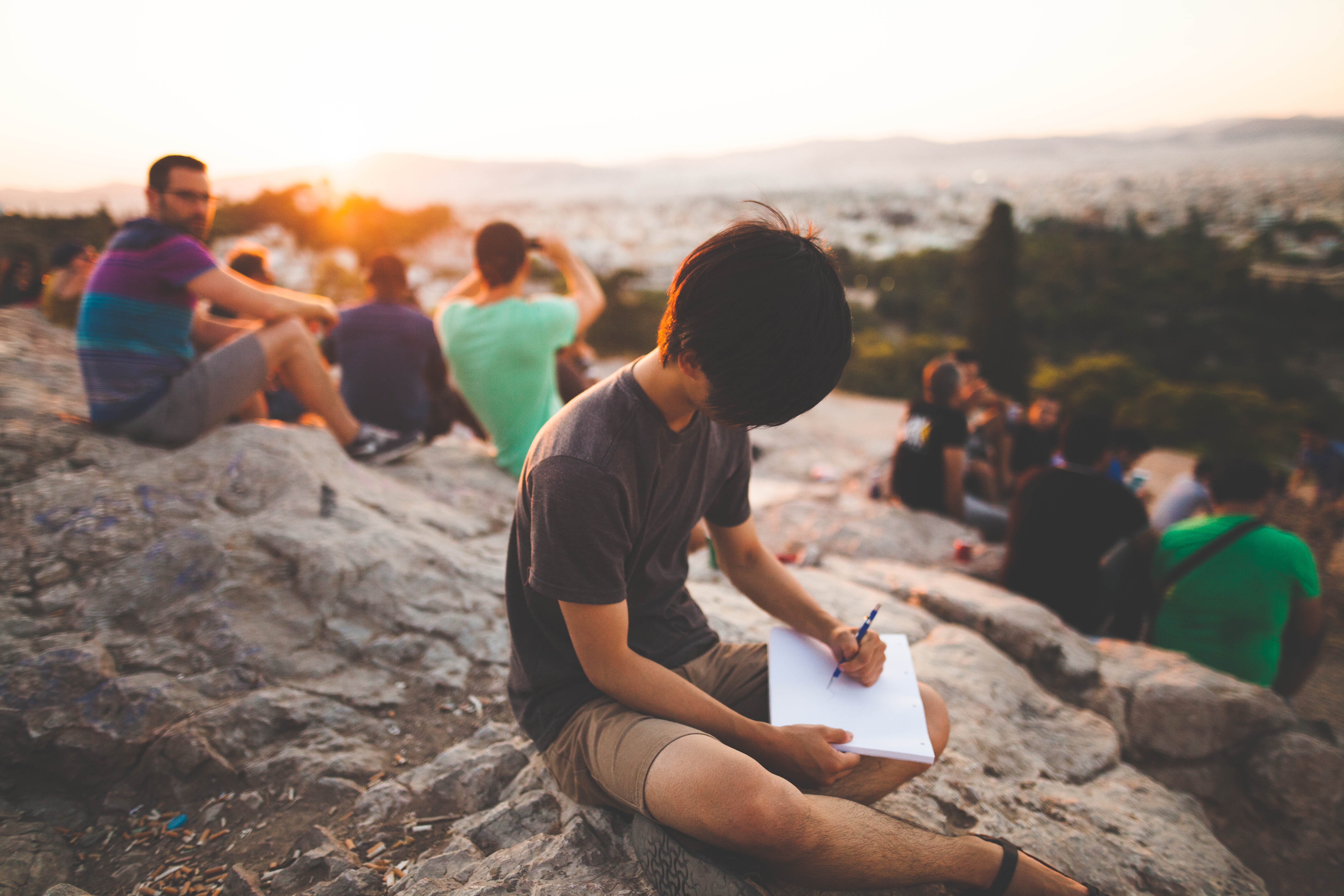 A teenager sits atop a rocky hill and writes into his journal. In the background, other teens are watching the golden sunset.